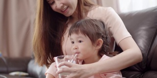 美丽的亚洲母亲给她的小女儿一杯牛奶。快乐可爱的蹒跚学步的女孩喝牛奶。
