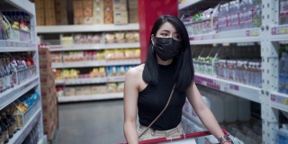 迷人的亚洲女人戴着防护面罩推着购物车在杂货店，货物工厂仓库得到供应，人类消费，covid19新常态，预算生活成本，超市过道行走