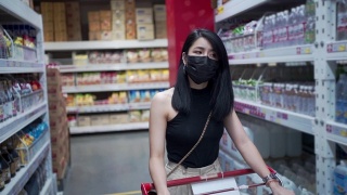 迷人的亚洲女人戴着防护面罩推着购物车在杂货店，货物工厂仓库得到供应，人类消费，covid19新常态，预算生活成本，超市过道行走视频素材模板下载