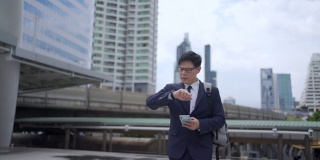 慢动作，微笑的年轻亚洲商人办公室职员穿着西装走在城市里，用智能手机发送在线信息。在市区使用手机工作或社交媒体的商人