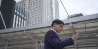 慢动作，微笑的年轻亚洲商人办公室职员穿着西装走在城市里，用智能手机发送在线信息。在市区使用手机工作或社交媒体的商人