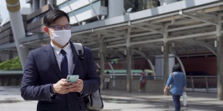 4K亚洲商务人士办公室人员走在城市火车站，使用智能手机和互联网为公司工作。商人戴口罩保护covid-19病毒大流行和空气污染。新标准的概念。