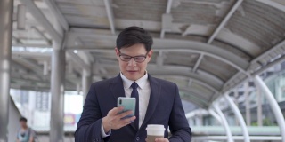 微笑的亚洲商人办公室职员拿着外卖咖啡杯走在城市里，用智能手机发送在线信息，在人群背景下进行户外企业工作。