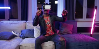 迷人的微笑年轻的黑人男子在虚拟现实耳机坐在沙发上的当代房间在晚上的时间