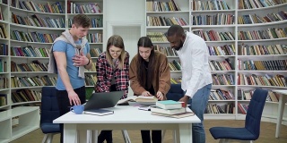 教育理念，四个满意的漂亮的多种族学生站在图书馆的桌子旁边看书，做笔记