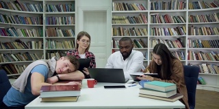 前视图疲惫的英俊男子，睡在桌子上和其他三个好看的多种族学生在图书馆的笔记本电脑上工作