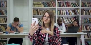 美丽微笑的现代明亮的女学生戴着眼镜坐在图书馆里，看着手机摄像头上其他人多样化的学生背景