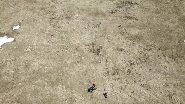 一对夫妇躺在田野上，在盛开的番红花中间。这些花生长在一片金黄色的干枯草地上。无人机飞回来了，让一切看起来更小了。冬天结束，春天开始