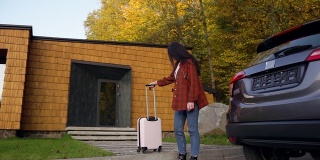 满意的年轻黑头发的女人，穿着休闲衣服，带着轮子上的手提箱到汽车前，她的旅行汽车