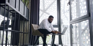 专注、睿智、蓄着胡须的非裔美国办公室职员，在一个有大窗户的现代办公室里，在电脑的另一个地方打字