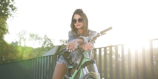 美丽的年轻女子在牛仔裤衣服和太阳镜坐在自行车上，摆姿势与真诚的微笑在日落在夏天。骑相机