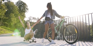 白种年轻女子坐在长板上，她最好的女性朋友骑着自行车走在她前面。享受生活