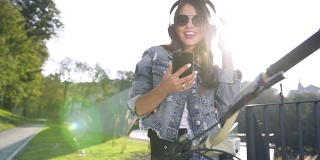 年轻女子戴着太阳镜穿着现代牛仔服装，一边骑自行车，一边通过手机上的应用程序在手持智能手机的无线耳机听音乐。日落