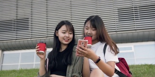 迷人的肖像两个愉快的亚洲褐发女人坐在草地上，使用他们的手机，喝咖啡在现代建筑的背景