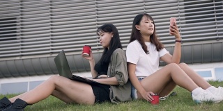 两个迷人的年轻亚洲女性最好的朋友背靠背坐在草坪上，一个在用手机，另一个在喝咖啡，在现代化的大楼附近用笔记本电脑工作