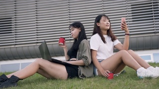 两个迷人的年轻亚洲女性最好的朋友背靠背坐在草坪上，一个在用手机，另一个在喝咖啡，在现代化的大楼附近用笔记本电脑工作视频素材模板下载