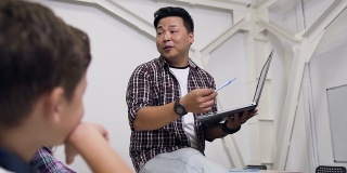 韩国老师坐在桌子上，手里拿着笔记本电脑给学生上课。小学生认真听讲，向老师提出重要问题