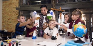 前视图。亚洲男性科学教师教年轻的学生做化学实验，拿着两个烧瓶液体引起强烈的蒸发