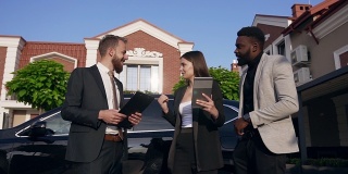 三个年轻的混合种族的办公室工作人员使用平板电脑和工人文件，站在美丽的汽车旁边的户外