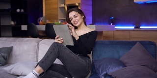 迷人的快乐的现代年轻褐发女性穿着黑色上衣和牛仔裤坐在家里的沙发上，使用着平板电脑