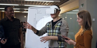 年轻的白人男子戴着虚拟现实眼镜玩游戏，而他的同事看着他
