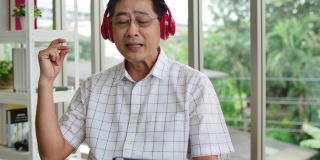 一个快乐的老人的肖像戴着耳机，拿着数字平板电脑听音乐在家里的客厅。