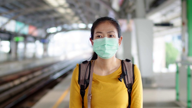 年轻的亚洲女子戴着口罩站在火车站的月台上。
