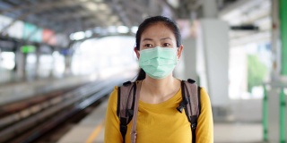 年轻的亚洲女子戴着口罩站在火车站的月台上。