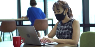 亚洲女员工戴口罩，在商务办公室使用笔记本电脑，与同事保持社交距离，以保护冠状病毒或covid-19疫情大流行。职场新常态政策