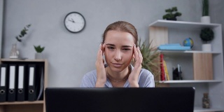 不开心的迷人的金发女孩按摩太阳穴，以缓解头痛时，她在现代橱柜的电脑