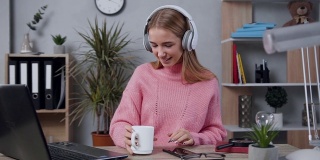 穿着针织毛衣的可爱快乐的年轻女子走到桌边，打开笔记本电脑，戴上耳机，享受着美妙的音乐