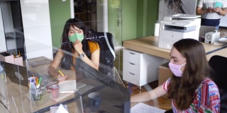 两个戴着口罩的商务人士在他们的办公桌前工作，他们之间有一个屏幕隔板