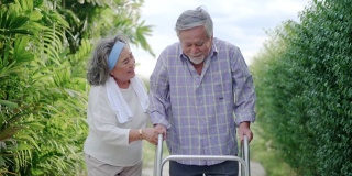 年老的亚洲妻子照顾她生病的丈夫使用帮助学习在房子周围的花园散步。老年保健与健康保险的概念