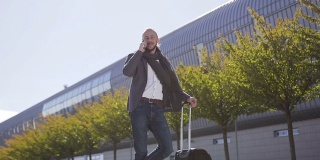 一个年轻的商人站在现代商务办公室附近，拿着行李箱在户外用智能手机说话。技术的概念，电话，商务旅行，商务，华尔街