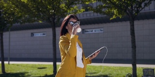 年轻漂亮的女商人戴着太阳镜，一边喝着咖啡一边在平板电脑上发信息。在户外。女学生在现代办公大楼附近使用平板电脑和喝咖啡