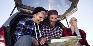 年轻的潮人朋友们坐在汽车后备箱里看地图，开心快乐。有趣的旅行和美丽的自然地点