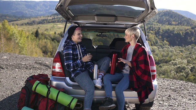 快乐的朋友是靠在后备箱车喝咖啡或茶在山的背景。两个快乐的时髦女孩坐在汽车后备箱里，喝着保温瓶里的热咖啡或茶，有说有笑。在户外