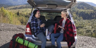 快乐的朋友是靠在后备箱车喝咖啡或茶在山的背景。两个快乐的时髦女孩坐在汽车后备箱里，喝着保温瓶里的热咖啡或茶，有说有笑。在户外