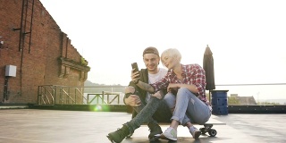 一对年轻帅气的夫妇正坐在现代建筑屋顶上的长板滑板上玩智能手机。两个年轻人在用智能手机自拍。现代城市生活方式的概念