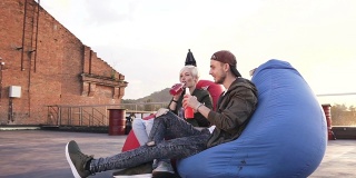 一对年轻迷人的夫妇有乐趣地坐在扶手椅袋喝美味的能量饮料在工业建筑的屋顶在日落。那个帅哥和他的女朋友在户外玩得很开心
