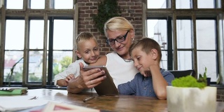 一位戴眼镜的中年白人商务妇女和她的孙子们坐在舒适的办公室里的桌子旁玩平板电脑游戏