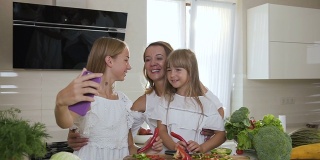 两个可爱的姐妹在厨房做饭的时候亲吻她们心爱的迷人的母亲。女孩们穿着白色衣服亲吻妈妈，在家里厨房用智能手机自拍