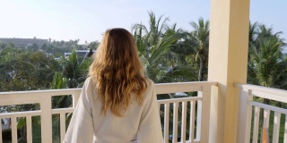 慢镜头:清晨，年轻女子拉开窗帘，走出酒店房间，走到阳台上。在豪华度假村度假的女人在早上打开窗帘，花园的景色