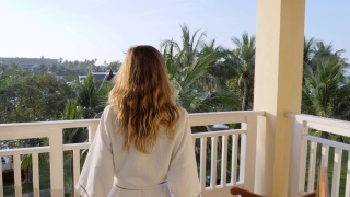 慢镜头:清晨，年轻女子拉开窗帘，走出酒店房间，走到阳台上。在豪华度假村度假的女人在早上打开窗帘，花园的景色视频素材模板下载