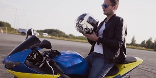 摩托车手穿着皮夹克和牛仔裤，走向他的摩托车，坐在他身上，戴着太阳镜