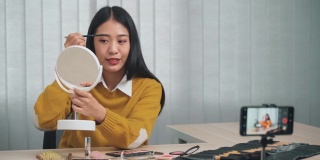 一名年轻的亚洲女子正在家里的视频博客中演示如何通过智能手机给社交媒体上的朋友化妆。