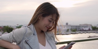 年轻美丽的亚洲女游客站在阳台上，用智能手机相机自拍或在日落时拍摄美丽的城市和河流。美丽的女孩放松，享受户外生活方式，度假旅行的城市。