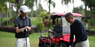 亚洲华人成年人从高尔夫球袋中挑选高尔夫球棒，并在高尔夫球场开球点指导教儿子