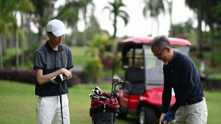 亚洲华人成年人从高尔夫球袋中挑选高尔夫球棒，并在高尔夫球场开球点指导教儿子视频素材模板下载