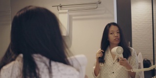 亚洲美丽的女人用吹风机梳理和造型她的头发镜子在酒店内部背景浴室。4 k慢动作。